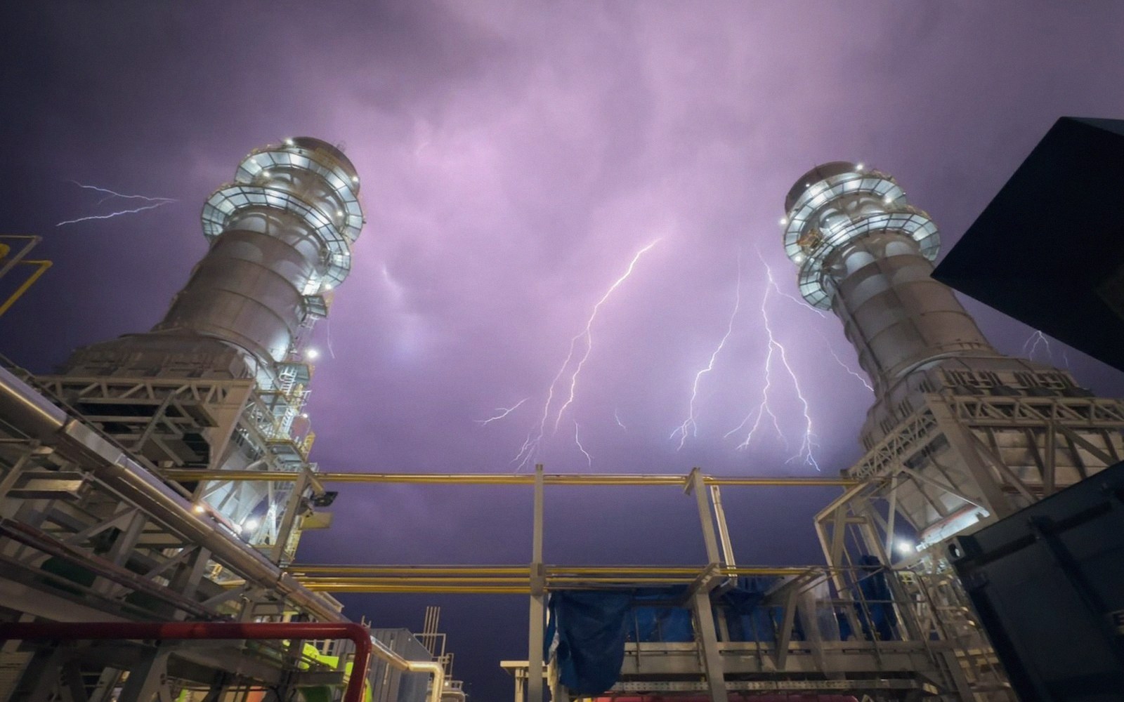 Lightning strikes behind two illuminated power plant smokestacks against a purple night sky