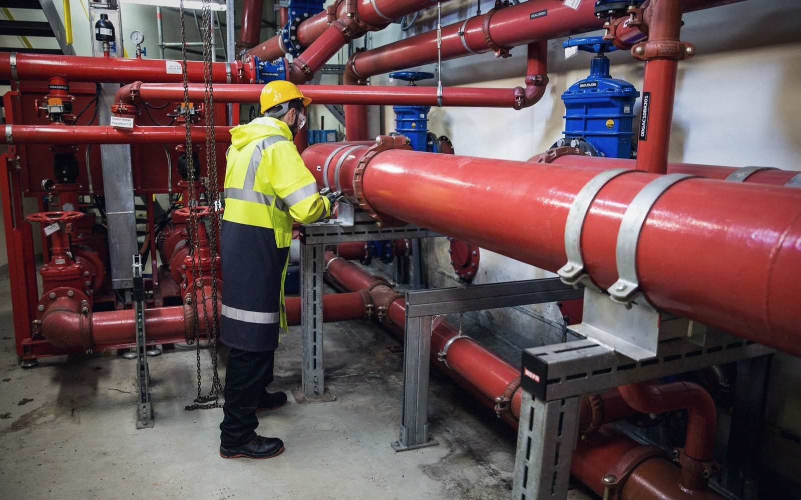 Worker in a hard hat inspects red pipework and blue valves in a mechanical room