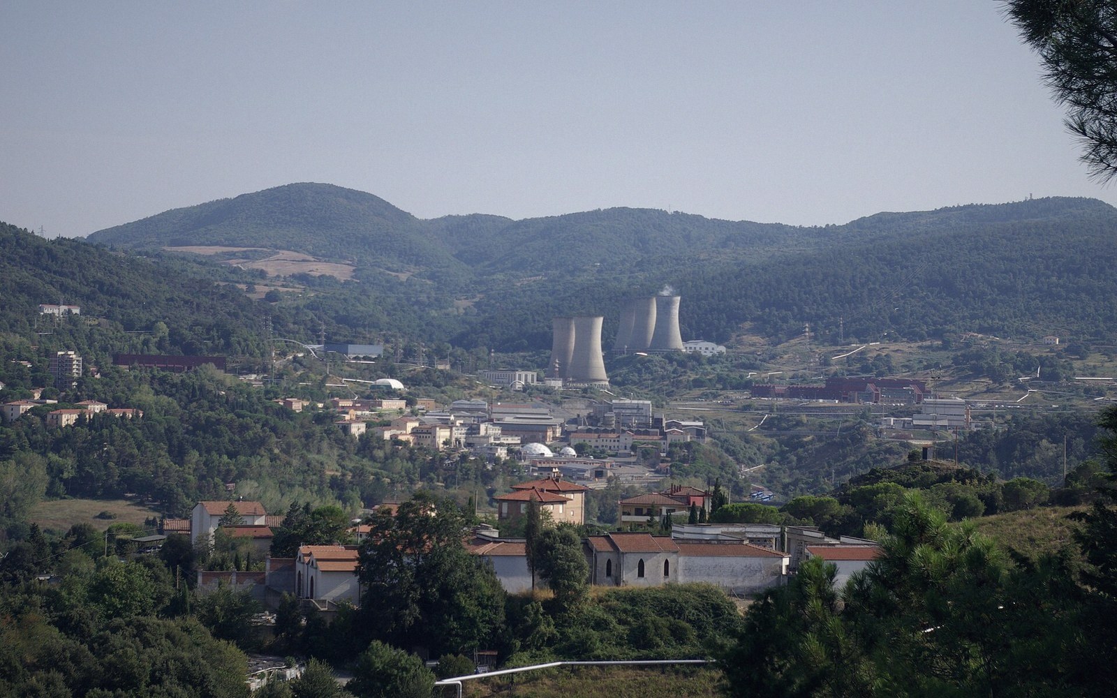 Cooling towers of a geothermal power plant rise above a hillside town surrounded by forested hills