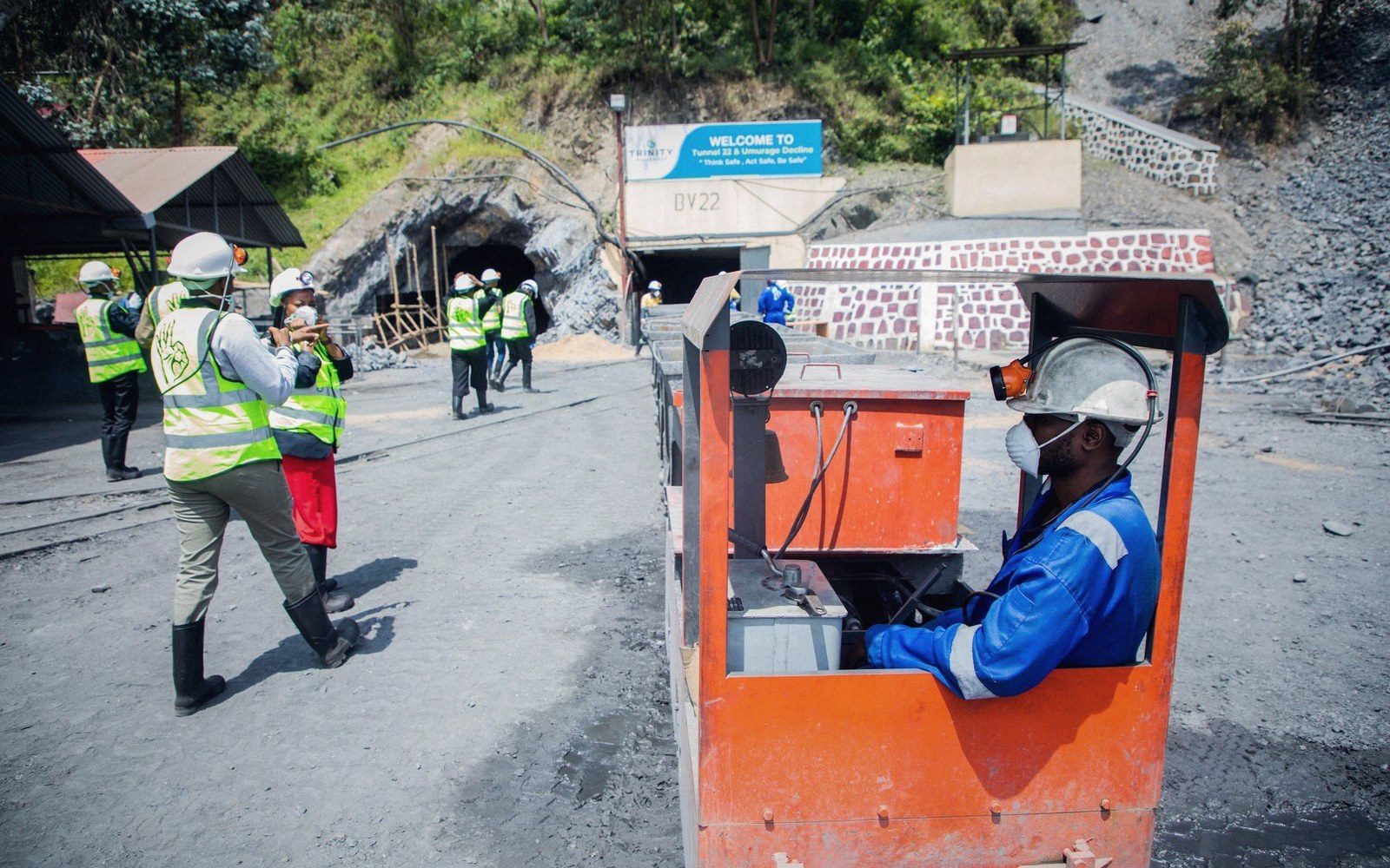 A miner operates an orange rail vehicle near a mine tunnel entrance as workers gather nearby