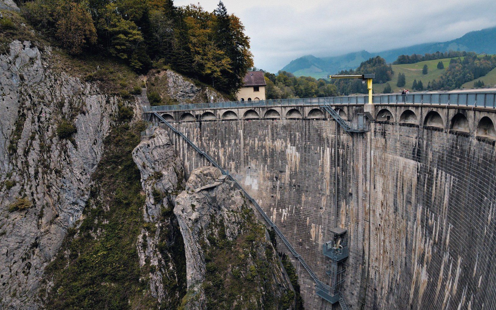 A tall arched dam wall curves along a rocky gorge beneath forested hills