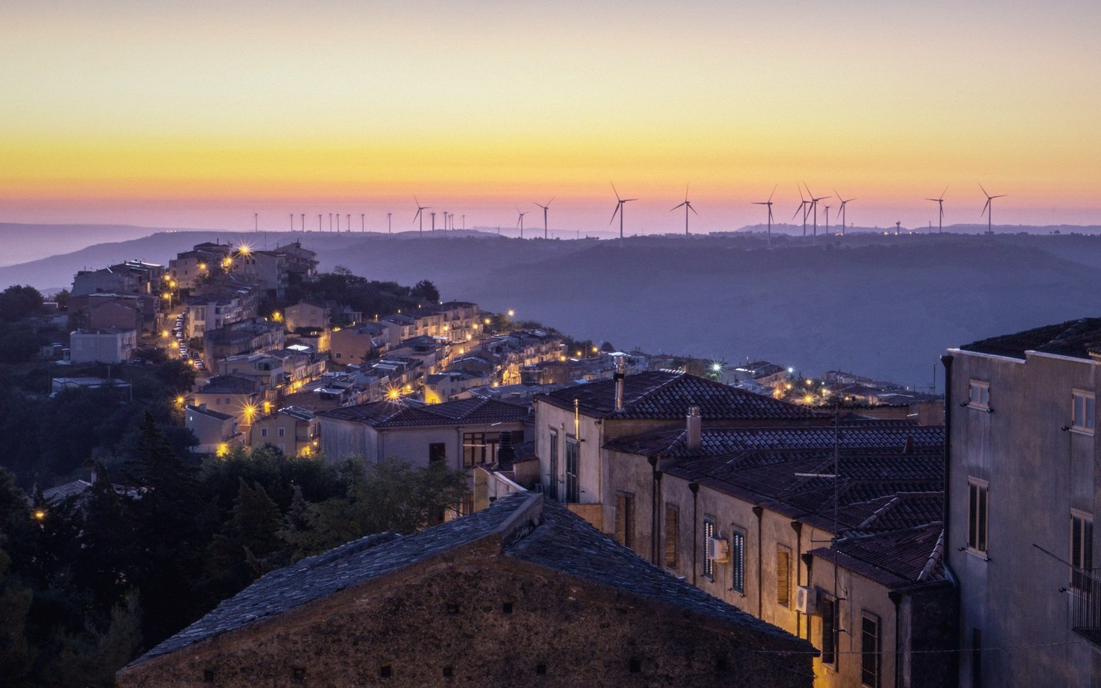 Hilltop town glowing at dusk with wind turbines lining distant ridges