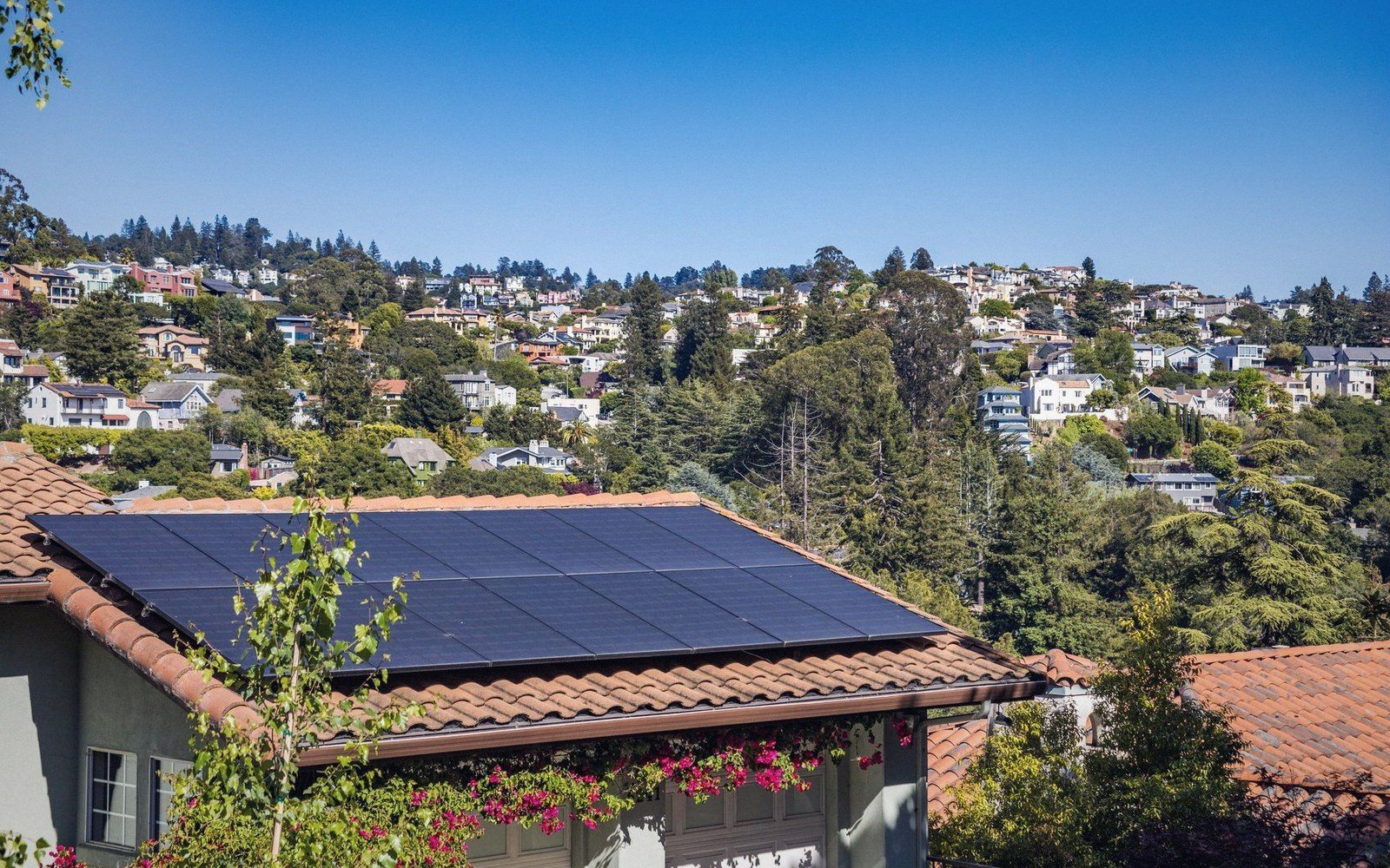Solar panels cover a tiled rooftop overlooking a hillside neighborhood of houses among trees