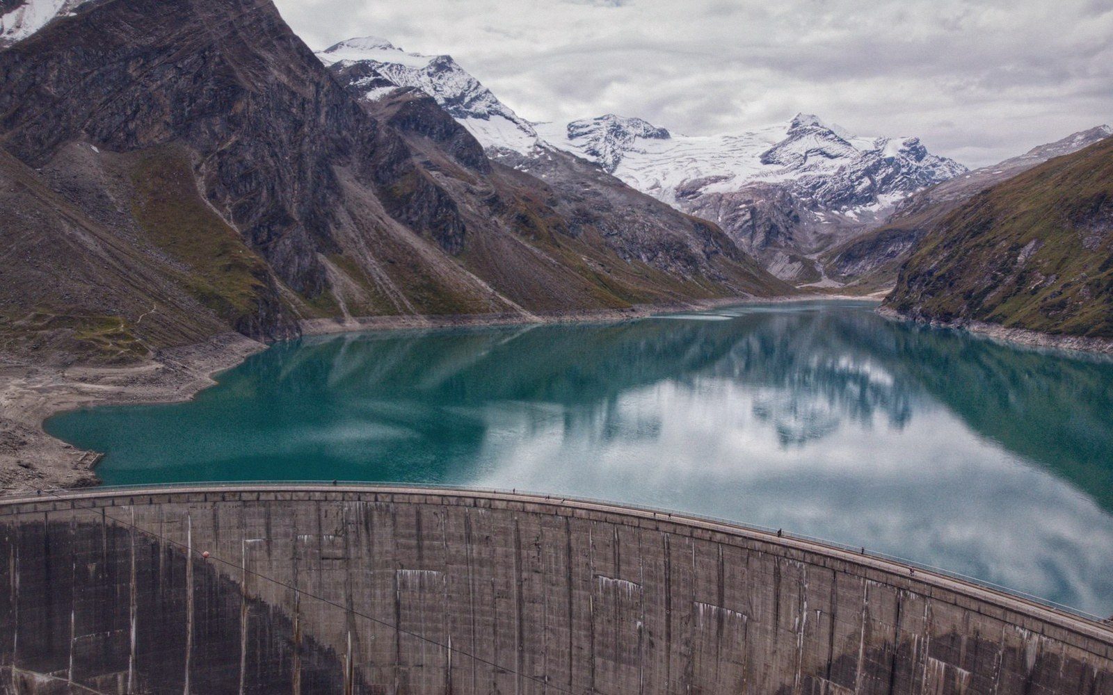 Curved concrete dam holds back a turquoise alpine reservoir surrounded by snow-capped peaks