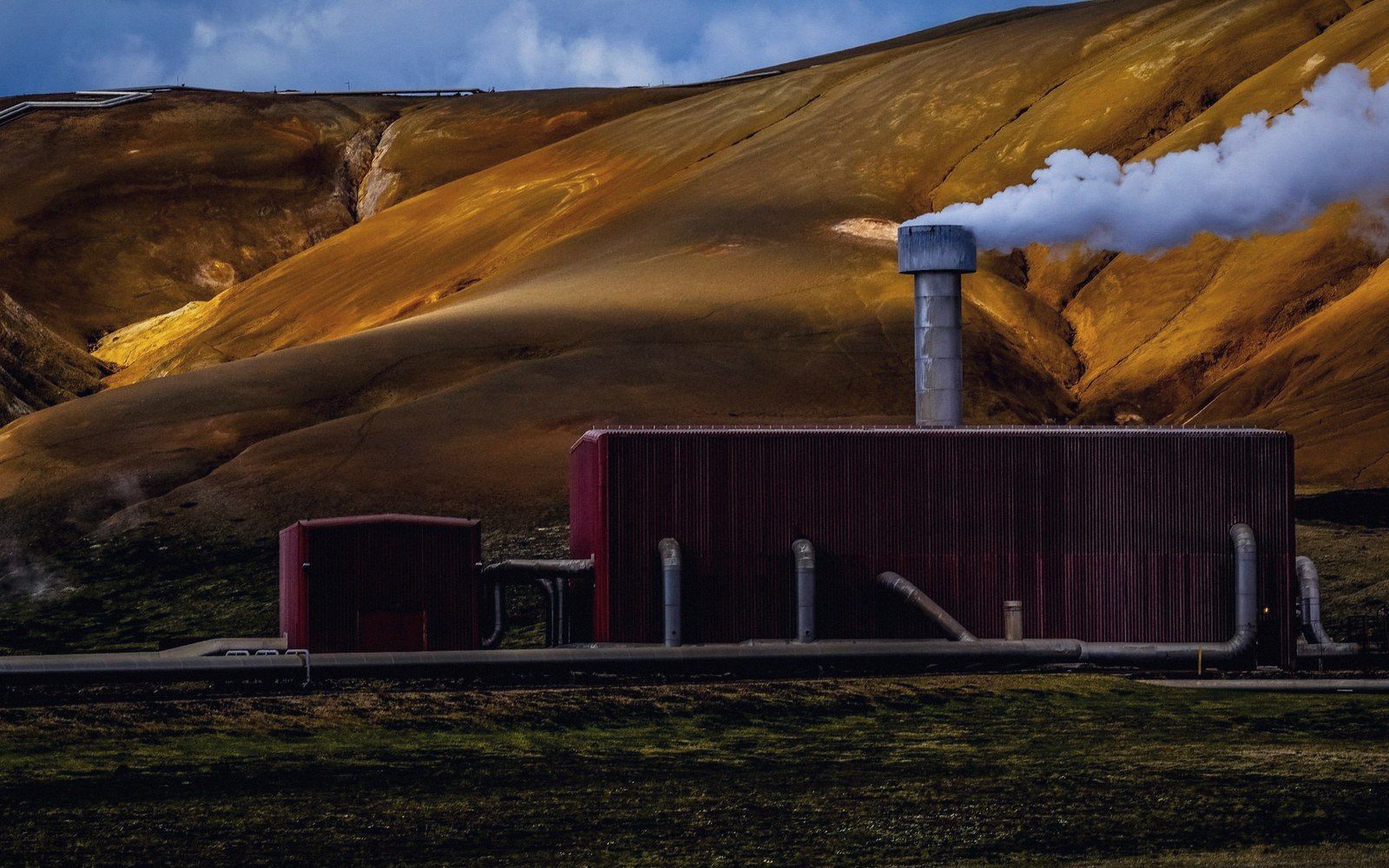 Steam vents from a geothermal power plant's chimney against a golden mineral-streaked hillside